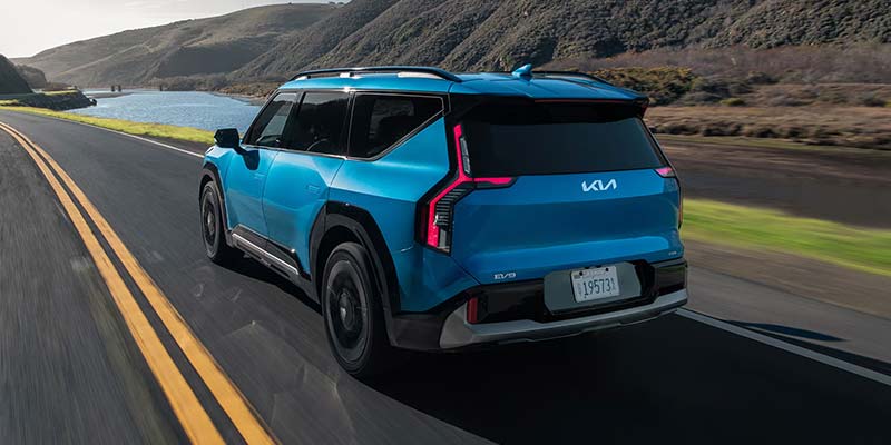 A teal Kia SUV drives towards the viewer on a coastal road overlooking mountains and the ocean