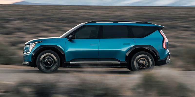 A vibrant blue Kia EV9 SUV, a modern electric vehicle, drives on a road with a blurred desert background