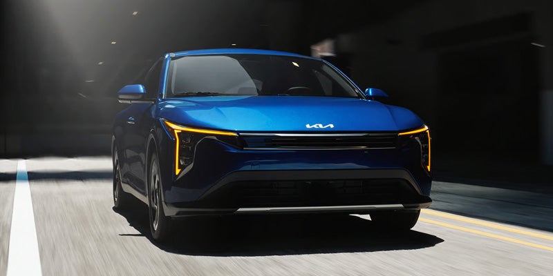 Front view of a blue Kia K4 sedan driving on a city street, featuring distinctive LED daytime running lights and a modern, aerodynamic design