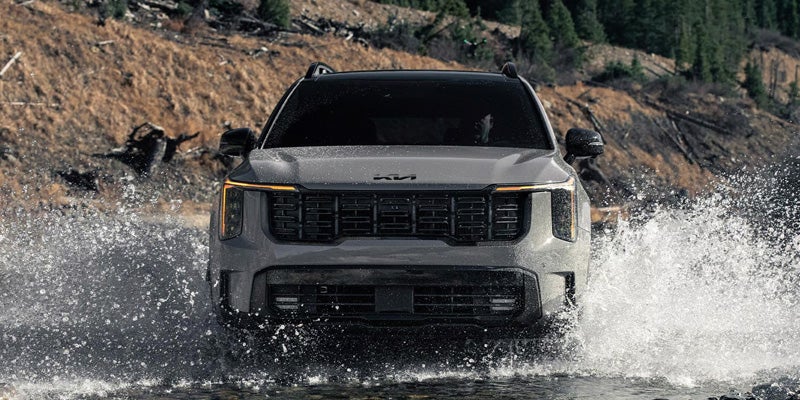 Front view of grey Kia Telluride SUV splashing through water, with a brown, treed embankment in the background