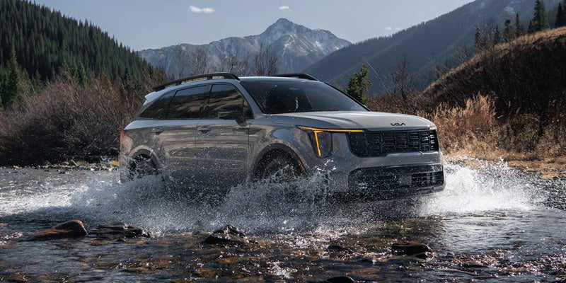 A silver Kia Telluride SUV splashing through a shallow river, with mountains and pine trees in the background