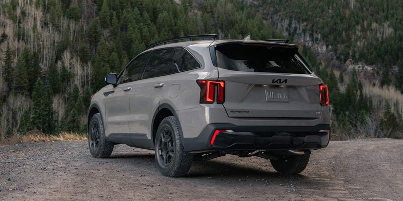 Rear view of a light gray Kia Sorento SUV with black wheels, parked on a dirt road in front of evergreen trees